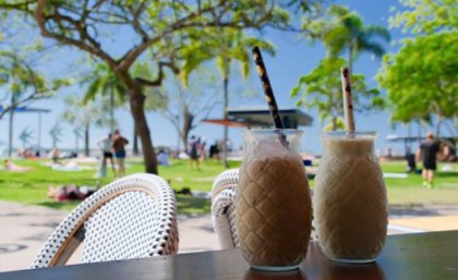 Two milky drinks with straws sit on a table in front of a green park with many people